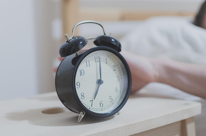 Photo of a black old style alarm clock and a hand leaning out of a bed to knock it off