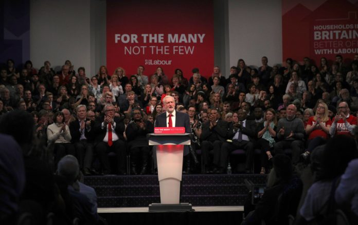 Jeremy Corbyn stands in front of a crowd at a conference with the sign behind reading 'For The Many, Not the Few'