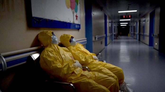 2 medical staff wearing yellow PPE and masks rest on chairs in a long hospital corridor.