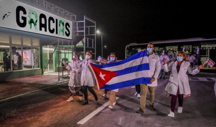 Doctors wearing white coats and masks walk along a road holding the Cuban national flag in front of a sign saying 