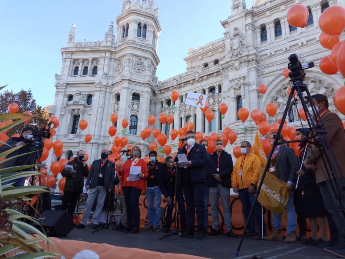 There is a platform in front of a fancy white building. Adults in masks holding orange balloons stand on the platform.