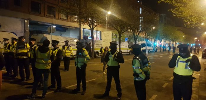 At night a line of Gardaí in high viz and Covid masks form a line across a street. There are lines of Garda vans in the background and a blurry image of a protester holding a placard.