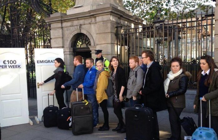 A number of young people queue in front of Leinster House with suitcases. They can choose between two doors, one of which says 