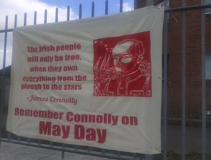 A white banner hangs up on a fence. It is hand painted in red. There is a picture of James Connolly and the words 